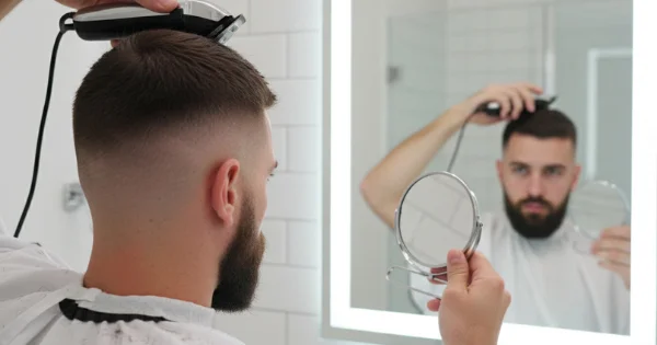 A man uses an electric clipper in front of a mirror to even out the top section, completing the final adjustments of a buzz cut fade.