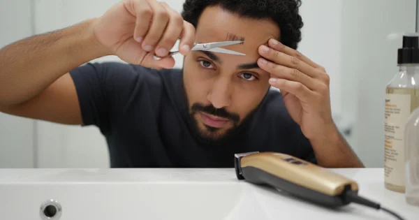 A man trims and cleans up his front hairline with small scissors, refining the edges to complete a neat buzz cut fade.